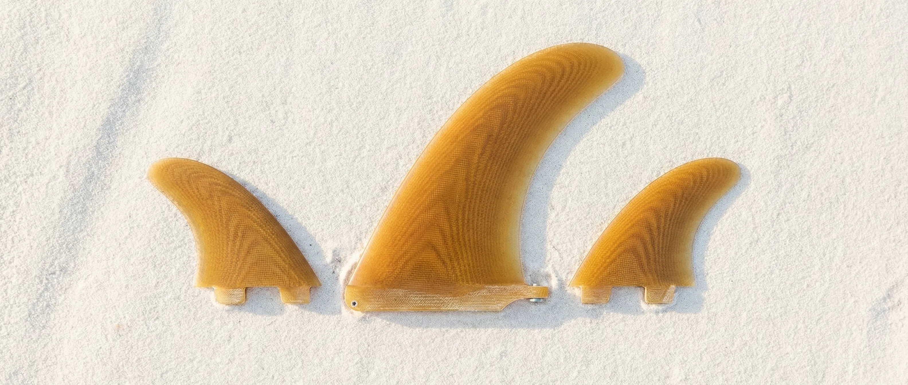 Three fiberglass surfboard fins in a 2+1 configuration laid out on white sand with the larger center fin flanked by two smaller side bites