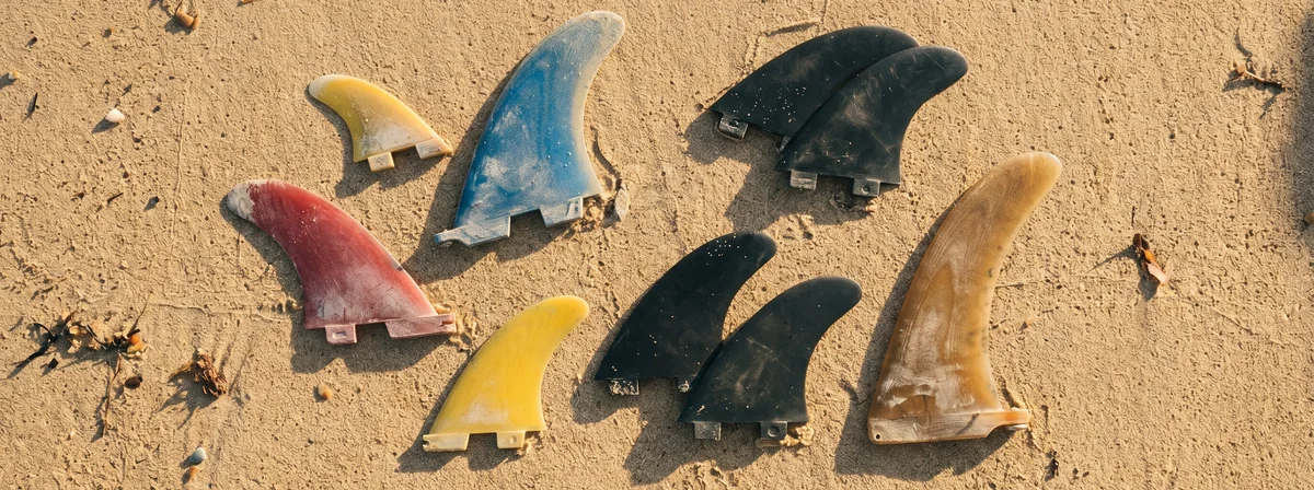 Overhead flat lay of assorted used surfboard fins arranged on golden beach sand at sunset, including thruster sets, twin keels, and a single longboard fin
