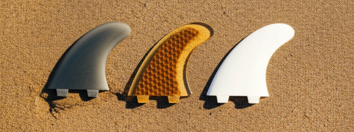 Three sets of surfboard fins laid on beach sand at golden hour showing different materials and constructions