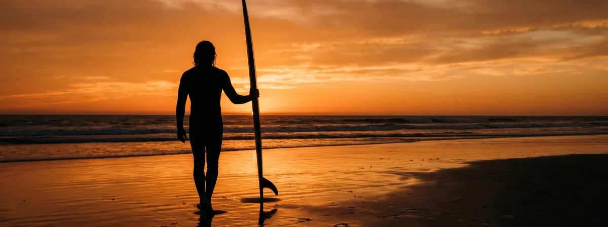 Silhouette of a surfer holding a surfboard at sunset, the board's bottom contour catching golden light along the rail