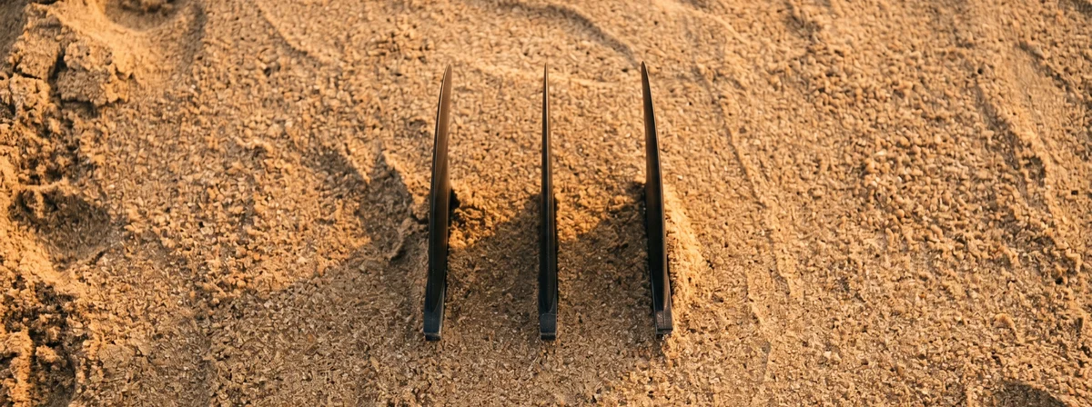 Three thruster fins in different materials laid out on warm beach sand at golden hour, showing fiberglass, carbon, and plastic construction side by side