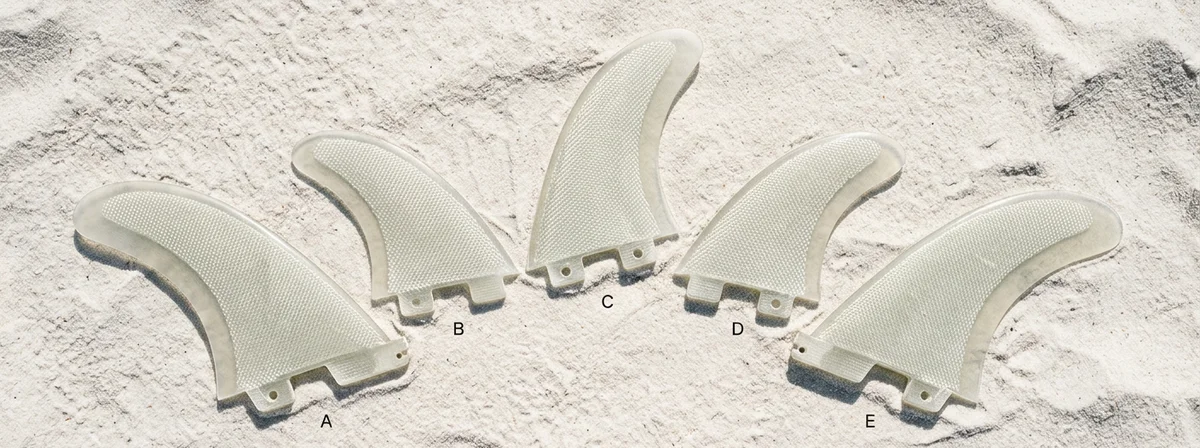 Five translucent fiberglass surfboard fins labeled A through E laid out on white sand showing a complete five-fin set