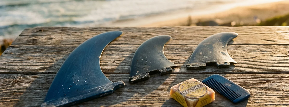 Three surfboard fins laid on a weathered wooden deck with surf wax and warm golden hour light