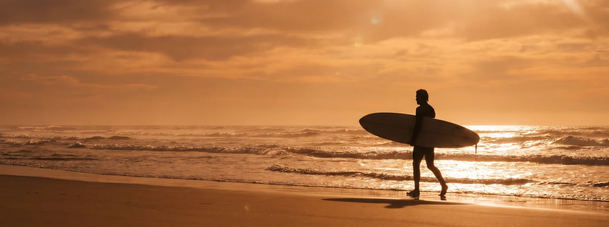 Surfer silhouetted at sunset holding a surfboard with a distinct tail shape against a backlit ocean
