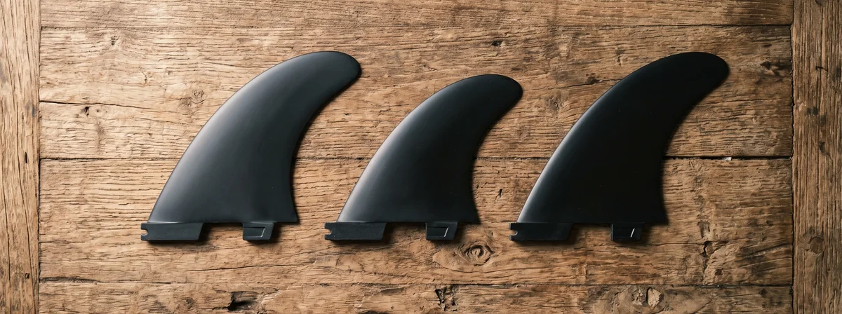 Three thruster surfboard fins laid out on a weathered wood table, with the center fin visibly smaller than the two side fins